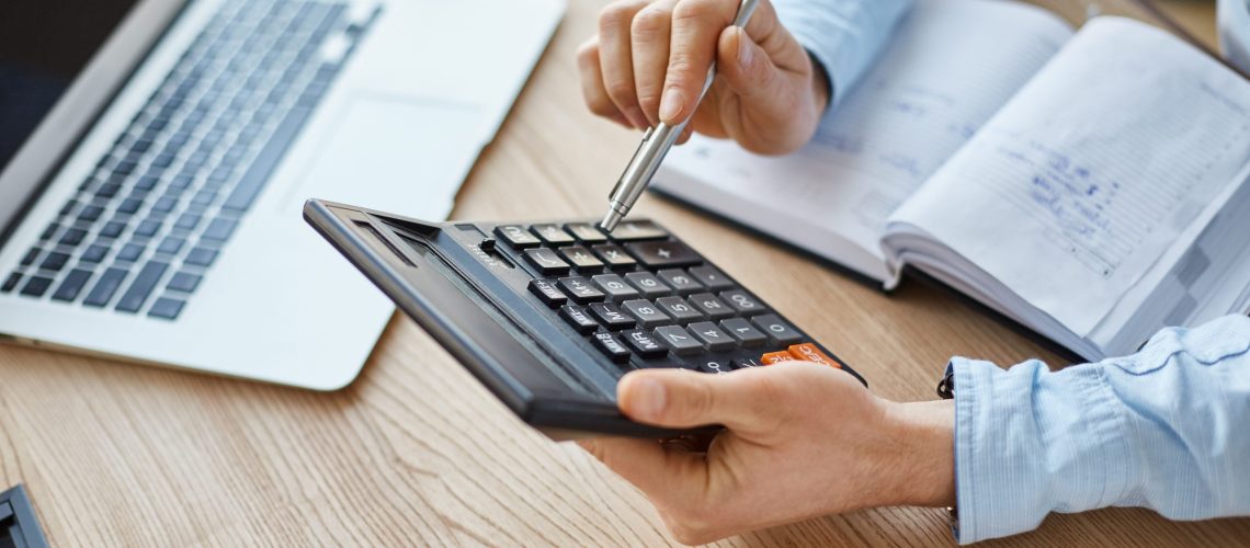 Cropped view of professional serious finance manager, holding calculator in hands, checking company month's profits, looking through details on laptop, writing down information for meeting with director
